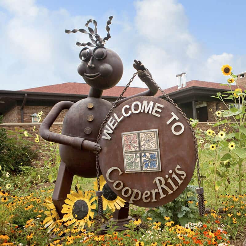 metal statue with curly hair holding welcome sign surrounded by blooming sunflowers