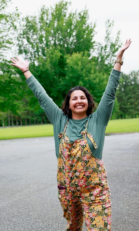 woman in overalls smiling with arms extended toward the sky