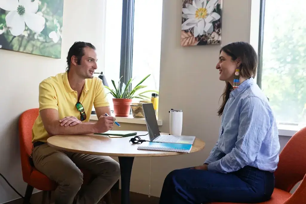 A young man and woman seated at a table with a laptop talk and laugh together