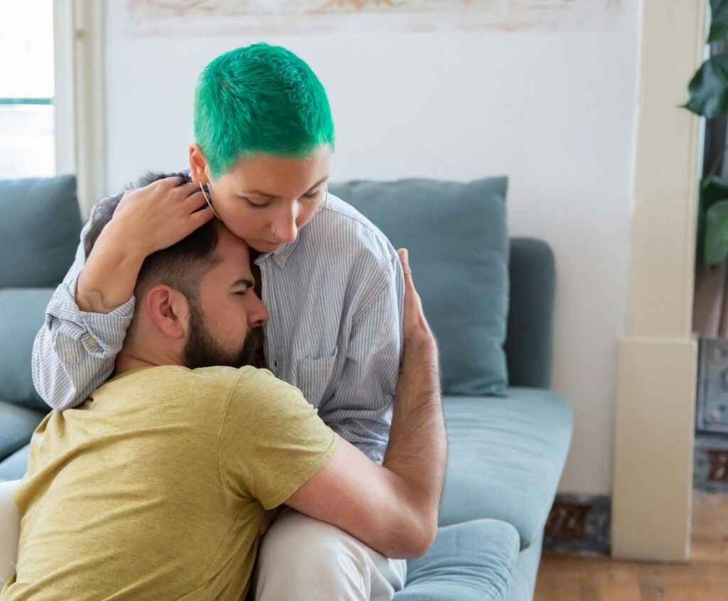 woman sits on a couch, hugging a man who sits on the floor, holding his head to her chest.. he looks distressed, she offers comfort.